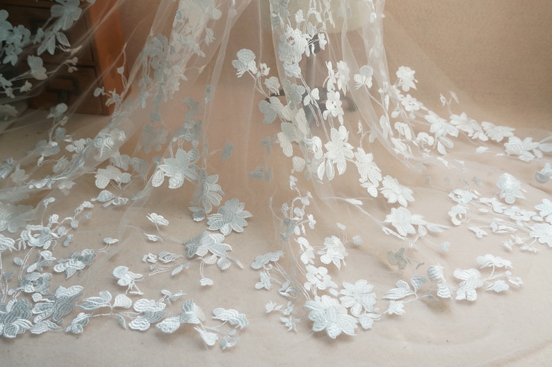 May include: Delicate white floral lace fabric. The lace features detailed, three-dimensional embroidered flowers and leaves. The fabric is semi-sheer, with a soft, flowing drape. Ideal for bridal wear, dresses, or decorative projects.