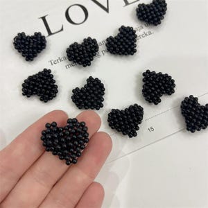 Può includere: Abbellimenti a forma di cuore neri con perline sparsi su una superficie bianca. I cuori sono fatti di piccole perline rotonde e sono di varie dimensioni. La parola "LOVE" è parzialmente visibile sullo sfondo.