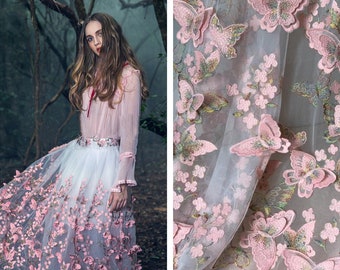 Può includere: Una gonna di tulle bianca con ricami di farfalle rosa. La gonna è a strati e ha un aspetto delicato e trasparente. La gonna è fotografata in un ambiente boschivo.
