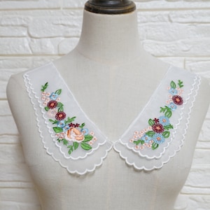 May include: White lace collar with scalloped edges and embroidered floral design in shades of pink, blue, and green.