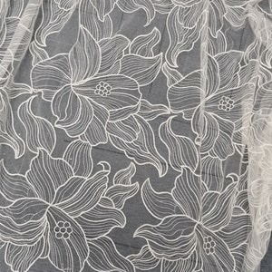May include: White floral lace fabric with a delicate, intricate design. The lace features a repeating pattern of stylized flowers with a textured, raised surface.