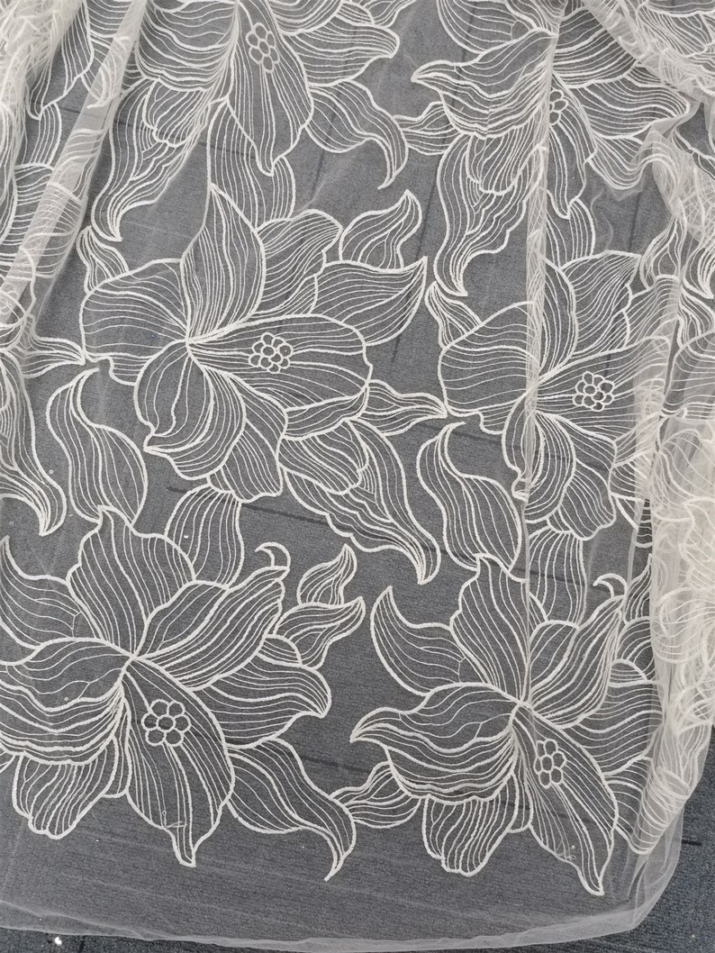 May include: White floral lace fabric with a delicate, intricate design. The lace features a repeating pattern of stylized flowers with a textured, raised surface.