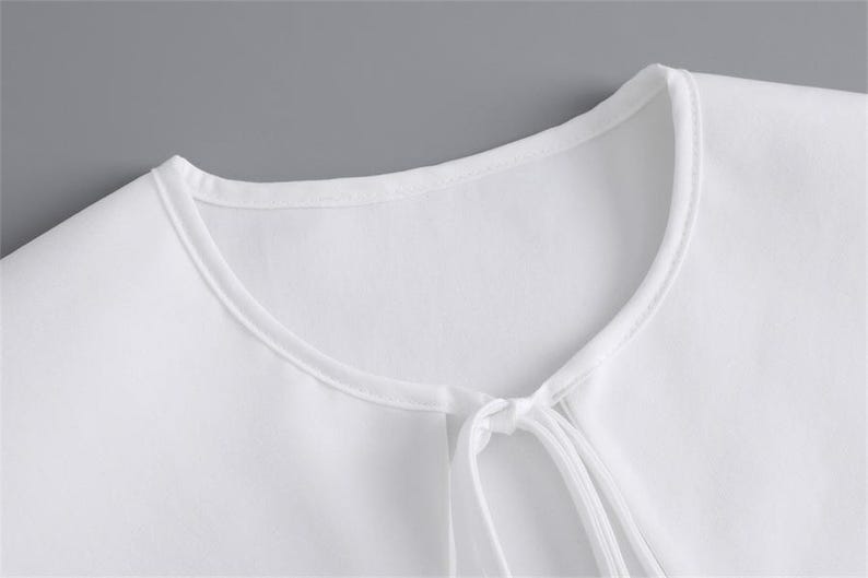 K&ouml;nnte beinhalten: Nahaufnahme eines wei&szlig;en Oberteils mit rundem Ausschnitt und Bindeband. Der Stoff scheint ein weiches, leichtes Material zu sein. Das Oberteil ist schlicht und einfach, mit minimalistischem Design.