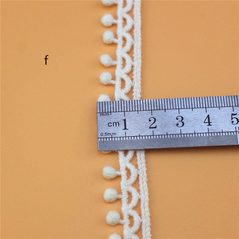 May include: White cotton lace trim with a scalloped edge and small pom-poms. The trim is measured with a ruler showing 1 cm.