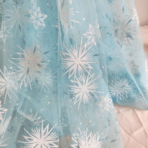 Pode incluir: Tecido de tule azul claro com um padrão de flocos de neve. O tecido apresenta grandes desenhos de flocos de neve brancos e pequenos detalhes de glitter prateado. O tule é drapeado, criando dobras suaves e uma aparência fluida. Ideal para fantasias ou projetos decorativos.