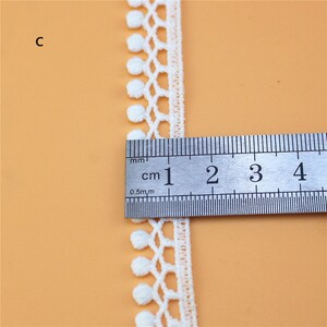 May include: White cotton lace trim with small pom-poms along the edge. The trim is measured with a ruler showing it is 1 cm wide.