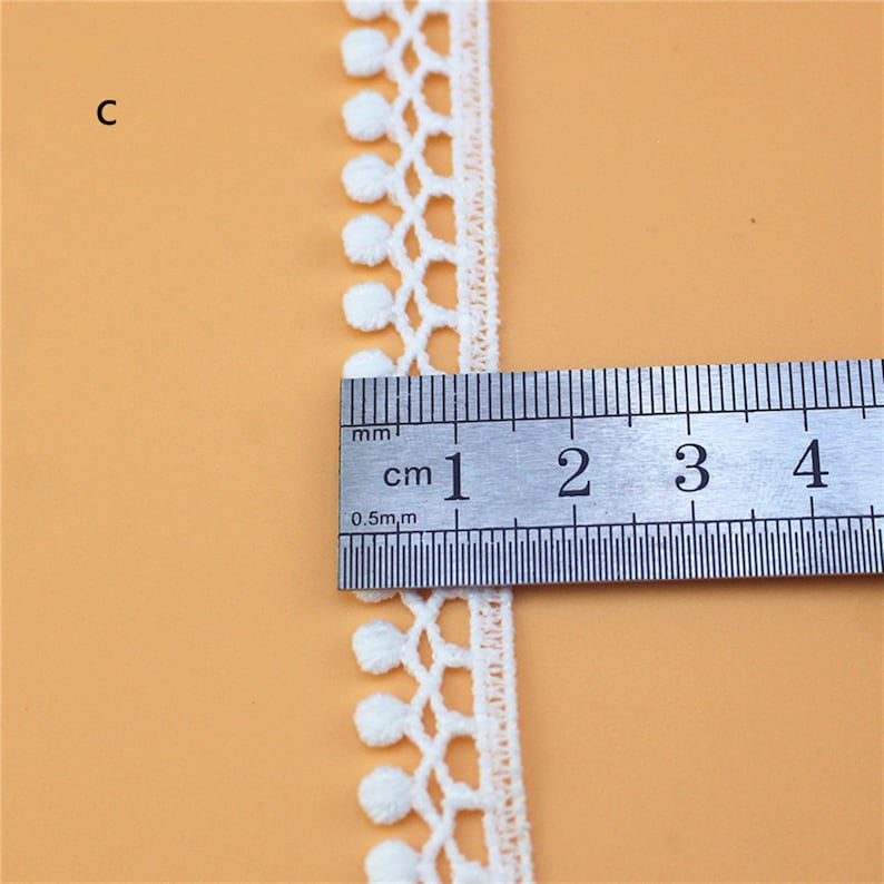 May include: White cotton lace trim with small pom-poms along the edge. The trim is measured with a ruler showing it is 1 cm wide.