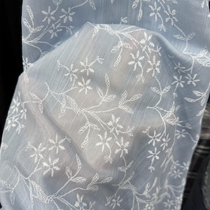 May include: A light blue, semi-sheer fabric featuring a delicate, repeating white embroidered pattern of small flowers and thin, winding leafy vines. The material has a subtle, fine vertical texture throughout, creating a light and airy appearance.
