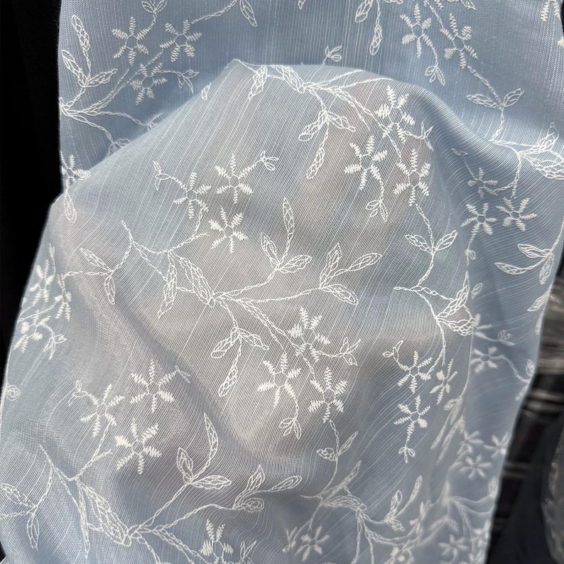 May include: A light blue, semi-sheer fabric featuring a delicate, repeating white embroidered pattern of small flowers and thin, winding leafy vines. The material has a subtle, fine vertical texture throughout, creating a light and airy appearance.