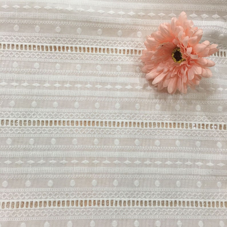 May include: White cotton fabric with a delicate embroidered pattern of circles, diamonds, and lines. The fabric has a sheer, airy feel and is perfect for creating garments or home decor projects.