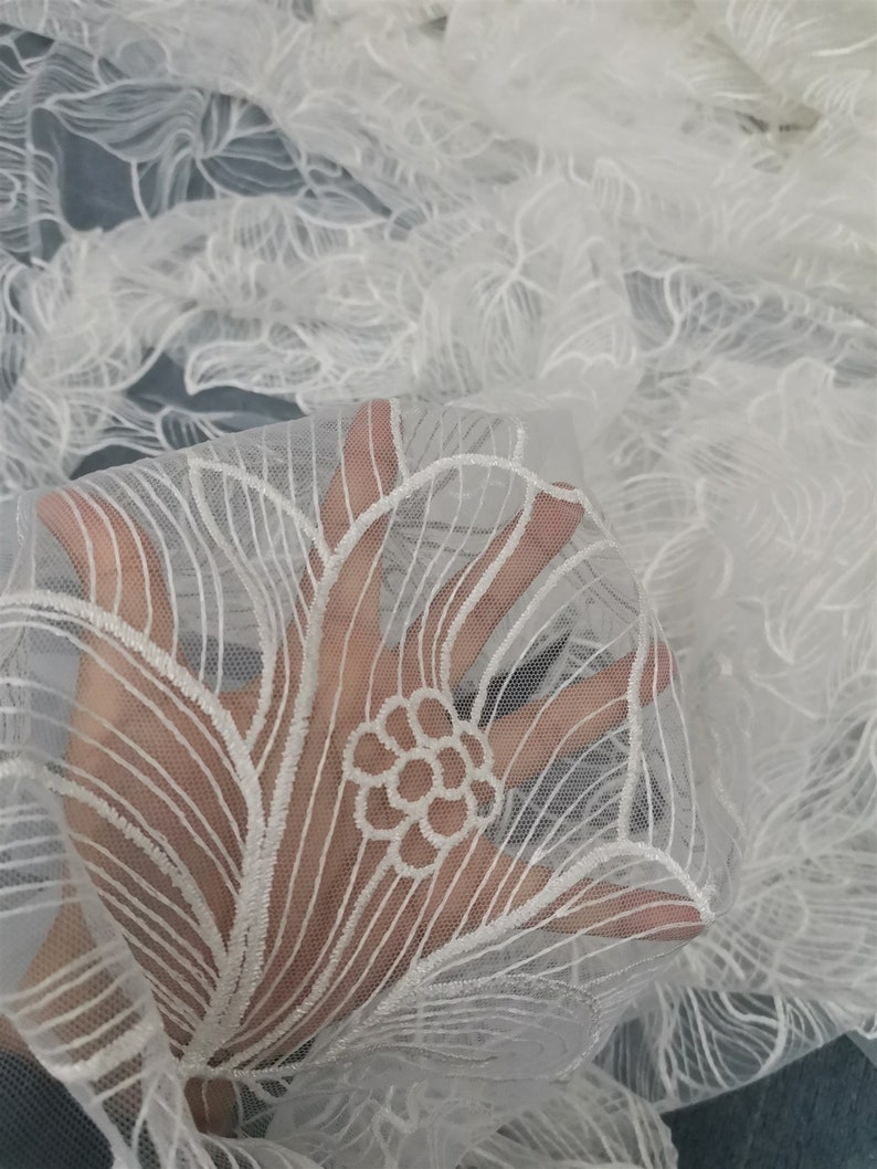 May include: White floral lace fabric with a delicate embroidered design. The lace is sheer and has a soft, flowing texture.