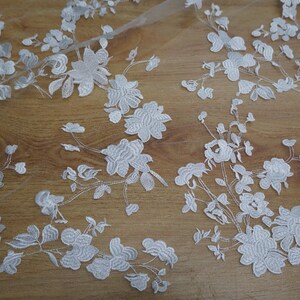 May include: White floral lace fabric with detailed embroidered flowers and leaves. The delicate design is set against a sheer background, creating a textured and elegant look. Ideal for bridal wear or decorative projects.