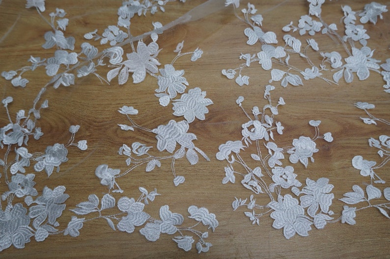 May include: White floral lace fabric with detailed embroidered flowers and leaves. The delicate design is set against a sheer background, creating a textured and elegant look. Ideal for bridal wear or decorative projects.