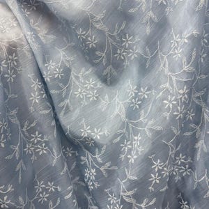 May include: A light blue fabric featuring a delicate white embroidered floral pattern with thin stems and small blossoms. The material has a subtle, semi-sheer quality with visible horizontal texture lines throughout the draped surface.
