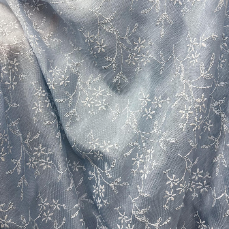 May include: A light blue fabric featuring a delicate white embroidered floral pattern with thin stems and small blossoms. The material has a subtle, semi-sheer quality with visible horizontal texture lines throughout the draped surface.