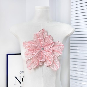 May include: A light pink floral applique with a central rhinestone detail, displayed on a white mannequin. The applique features multiple layers of petals and leaves, embellished with sequins. The design is delicate and feminine.