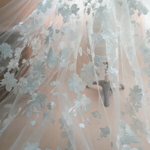 May include: Delicate white floral lace fabric. The fabric features an intricate pattern of embroidered flowers and leaves. The lace is semi-sheer, allowing light to pass through, and is likely used for bridal wear or other special occasion garments.