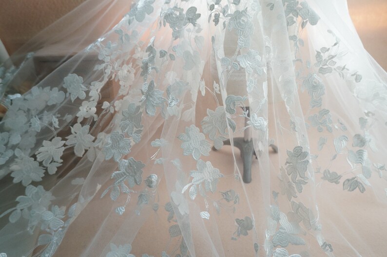 May include: Delicate white floral lace fabric. The fabric features an intricate pattern of embroidered flowers and leaves. The lace is semi-sheer, allowing light to pass through, and is likely used for bridal wear or other special occasion garments.
