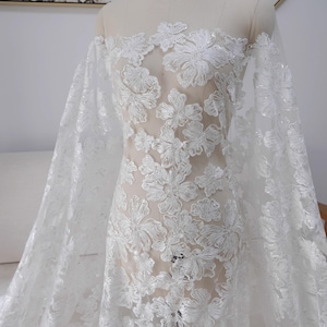 Può includere: Tessuto di pizzo floreale bianco con una finitura delicata e scintillante. Il pizzo presenta grandi motivi floreali intricati con un bagliore sottile.