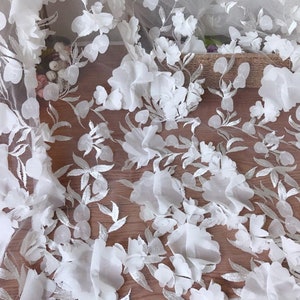 May include: White floral lace fabric with 3D flowers. The fabric has a delicate, sheer texture and is perfect for adding a touch of elegance to any project.