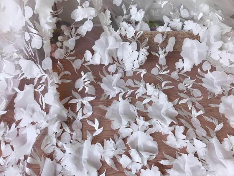 May include: White floral lace fabric with 3D flowers. The fabric has a delicate, sheer texture and is perfect for adding a touch of elegance to any project.