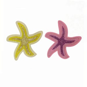 Peut inclure: Deux écussons en forme d'étoile de mer ornés de perles complexes. L'un est jaune avec des perles dorées, tandis que l'autre est rose avec des perles violet foncé. Les deux présentent un design à cinq branches avec un détail central circulaire.
