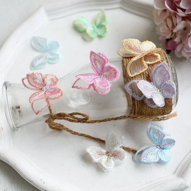 May include: Assortment of colorful butterfly-shaped fabric appliques with iridescent shimmer. The appliques are arranged on a glass jar and a white surface.
