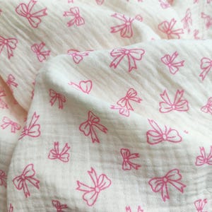 Könnte beinhalten: Cremefarbener Stoff mit einem sich wiederholenden Muster aus rosa Schleifen. Die Schleifen sind umrandet und über dem weichen, strukturierten Material verstreut. Der Stoff ist gefaltet und erzeugt Schatten und Tiefe.