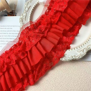 May include: Close-up of a vibrant red lace trim with multiple layers of ruffled fabric. The trim features a delicate floral lace edge and a sheer mesh backing, perfect for crafting and sewing projects.