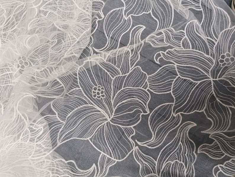 May include: White floral lace fabric with a delicate, intricate design. The lace is sheer and has a soft, flowing texture.