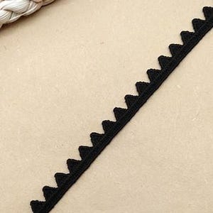 May include: Black decorative trim with a scalloped edge. The trim is made of a woven material and is angled across a tan surface. The trim is approximately 1.3 cm wide.