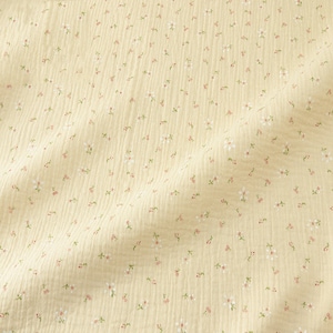 May include: A close-up of a light yellow fabric with a delicate floral pattern. Tiny white flowers with red centers and green leaves are scattered across the textured surface, creating a soft, feminine aesthetic. The fabric has a crinkled appearance.