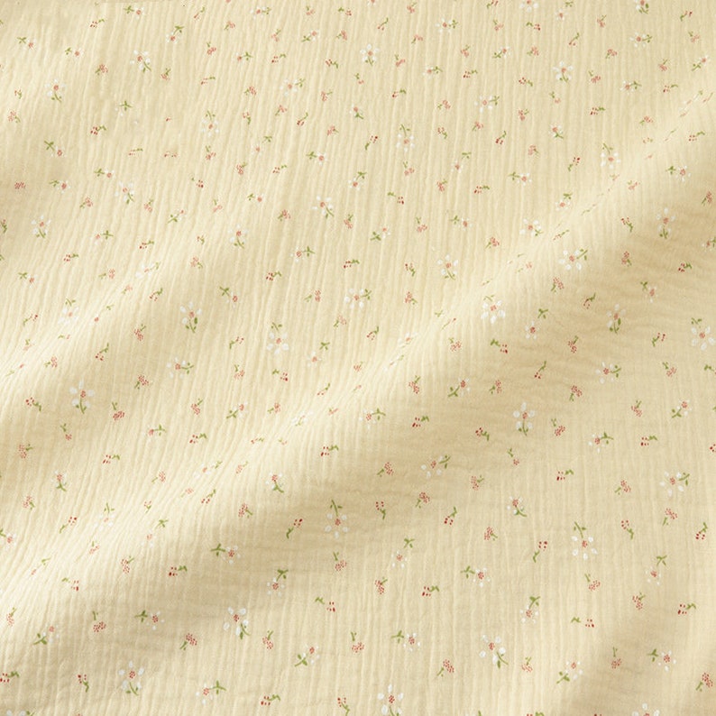 May include: A close-up of a light yellow fabric with a delicate floral pattern. Tiny white flowers with red centers and green leaves are scattered across the textured surface, creating a soft, feminine aesthetic. The fabric has a crinkled appearance.