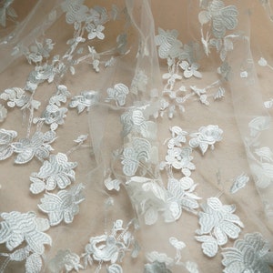 May include: Close-up of white floral lace fabric. The delicate lace features embroidered flowers and leaves, creating a textured pattern. The fabric is semi-sheer, with a soft, flowing drape. Ideal for bridal wear or decorative projects.