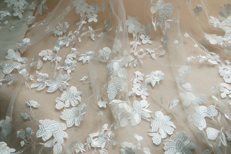 May include: Close-up of white floral lace fabric. The delicate lace features embroidered flowers and leaves, creating a textured pattern. The fabric is semi-sheer, with a soft, flowing drape. Ideal for bridal wear or decorative projects.