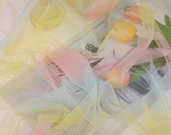 May include: A rainbow ombre tulle fabric with a delicate texture. The fabric is draped over a book and a few tulips.