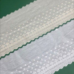 Könnte beinhalten: Weiße Baumwollspitzenborte mit einem gewellten Rand und einem zarten Blumenmuster. Die Spitze ist in der Mitte gefaltet, wodurch die filigranen Details des Designs sichtbar werden.