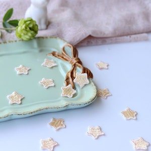 May include: A light green ceramic tray with a gold ribbon and a scattering of small, gold star-shaped embellishments. The tray is on a white surface with more gold star embellishments scattered around it.