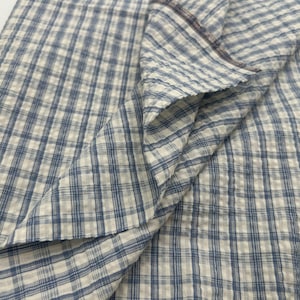 May include: A piece of fabric featuring a blue and white checkered pattern. The material has a visible seersucker texture with a puckered surface, creating a grid of small squares throughout the textile. The fabric is folded to show depth.