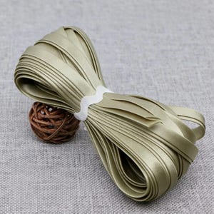 Può includere: Un rotolo di nastro di raso verde salvia fissato con una fettuccia bianca a zig-zag. Il nastro ha una finitura liscia e riflettente ed è avvolto a forma di otto. Una piccola sfera rotonda in rattan intrecciato marrone è posta sotto.