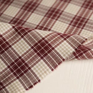May include: Close-up of a fabric with a plaid pattern in burgundy and beige. The fabric is folded, revealing the texture and weave of the material. The plaid design features intersecting lines creating a classic, timeless look. The fabric is likely used for apparel or home decor.