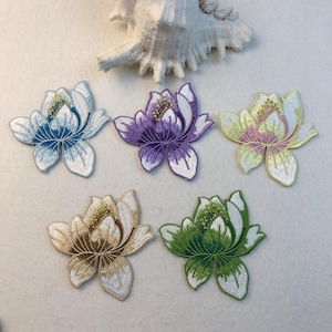 Può includere: Cinque toppe floreali ricamate in vari colori: blu, viola, giallo-rosa, marrone-oro e verde. Ogni toppa presenta cuciture dettagliate e un bordo bianco, che ricorda i fiori di loto. Sullo sfondo c'è una conchiglia.