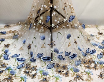 May include: Beige tulle fabric with blue and gold embroidered flowers. The fabric is sheer and delicate, with a floral pattern.