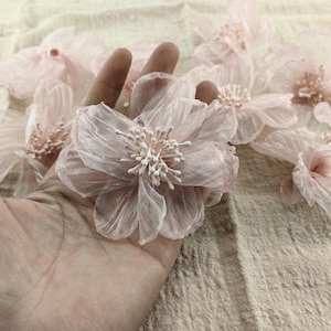 Pode incluir: Uma coleção de delicadas flores de tecido rosa claro e transparentes com centros brancos. As flores estão dispostas em uma mão, com a maior flor no centro.
