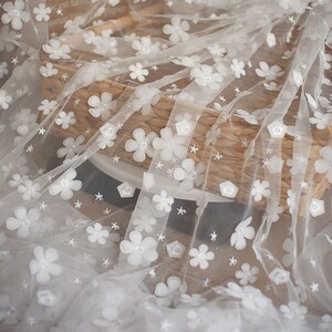 May include: White tulle fabric with white embroidered flowers and stars. The fabric is draped over a woven basket.