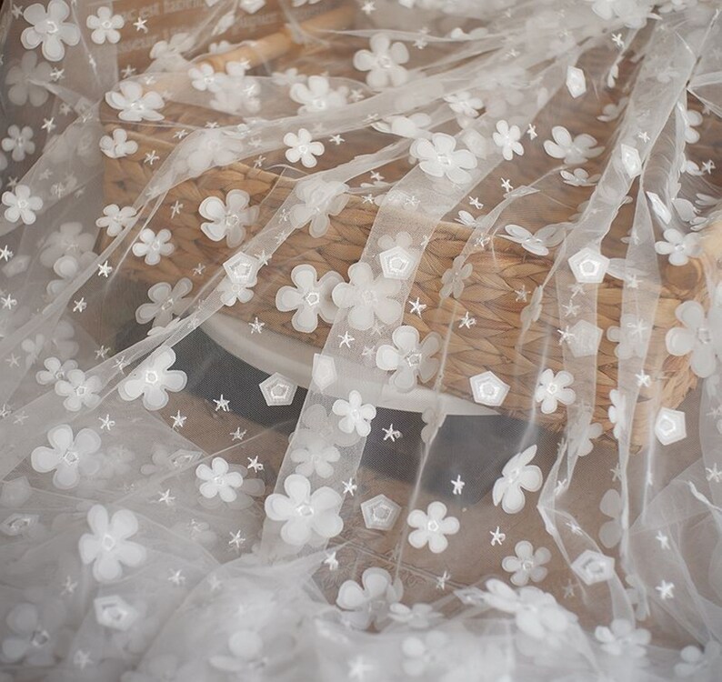 May include: White tulle fabric with white embroidered flowers and stars. The fabric is draped over a woven basket.