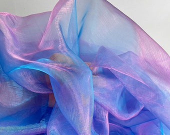 May include: A flowing fabric in shades of blue, purple, and pink. The sheer material has a subtle sheen, creating a shimmering effect. The fabric is draped and folded, showcasing its texture and color variations. Ideal for apparel or decorative purposes.
