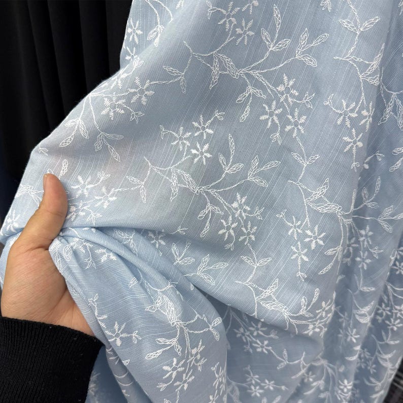 May include: A light blue fabric featuring a delicate white embroidered floral pattern with thin stems and small star-shaped blossoms. The material has a subtle vertical texture and a semi-sheer quality, showcasing intricate needlework throughout.