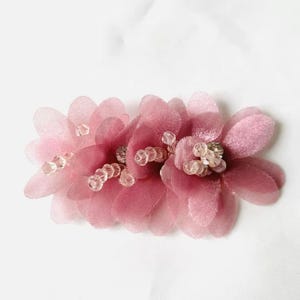 Pode incluir: Um acessório de cabelo com múltiplas pétalas de tecido sobrepostas em tons de rosa seco e malva. O centro do arranjo floral é acentuado com pequenas contas transparentes que adicionam um brilho subtil às pétalas de tecido macio.