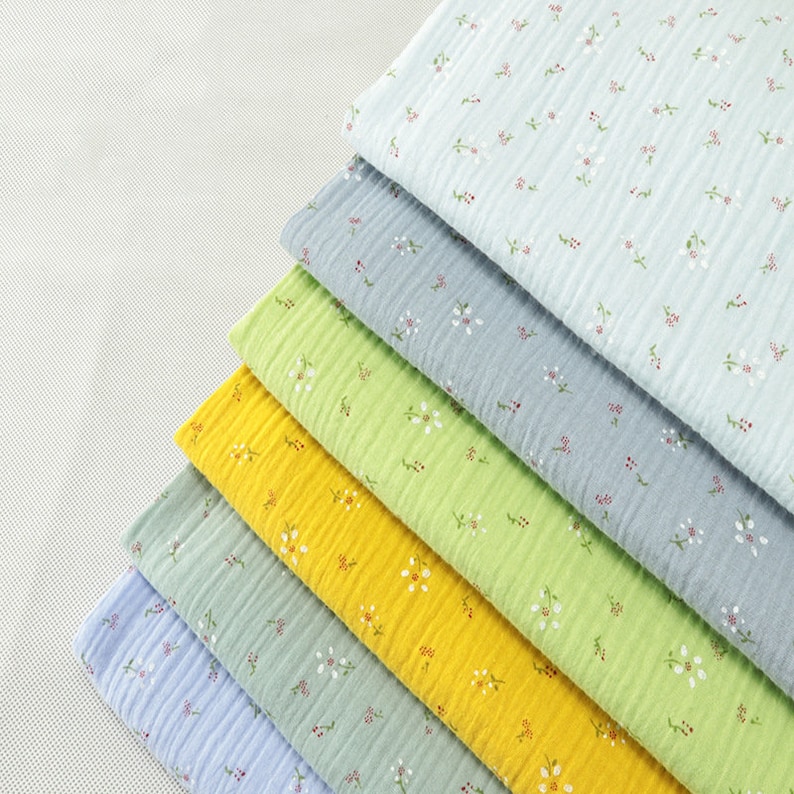 May include: A stack of colorful cotton fabric with a crinkled texture. The fabric is in shades of blue, green, and yellow, each adorned with a delicate floral pattern in white and red. The layers are neatly arranged, showcasing the variety of colors and designs.